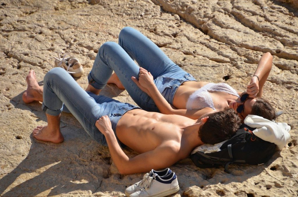 Couple relaxing on rocky beach in casual summer attire, enjoying the sun's warmth while lying close together
